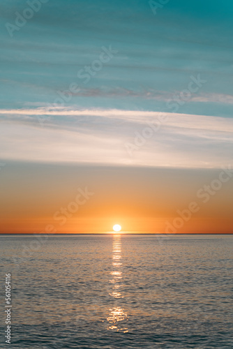 An amazing sunset with bright orange hues against a blue cloudless sky. A ray of sun casts a bright glare on the calm water surface of the ocean. The perfect evening in nature. © Pavel