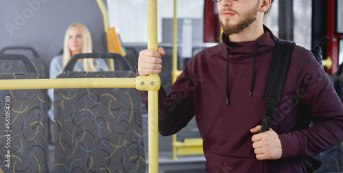 Close up of young man with beard wearing khudi, standing on bus, holding backpack, looking forward. Adult brunette traveling by public transport, busy. Concept of urban life.