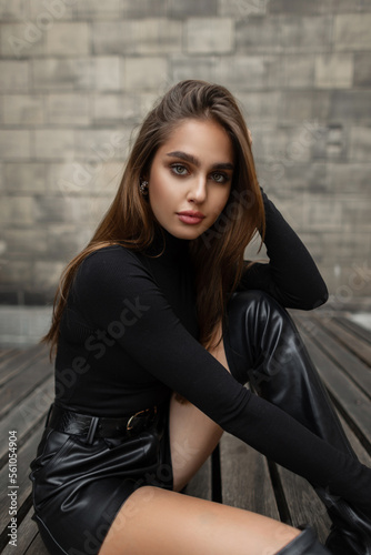Stylish beautiful young woman model in fashion black clothes with a black long sleeve shirt and leather shorts with boots sits on a wooden bench. Pretty elegant girl
