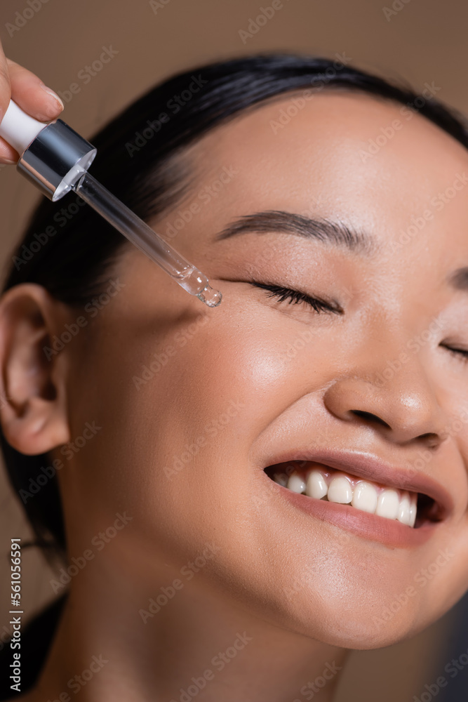 Close up view of smiling asian woman applying cosmetic serum on face isolated on brown.