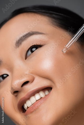 close up view of smiling asian woman applying cosmetic serum during facial pampering isolated on grey.