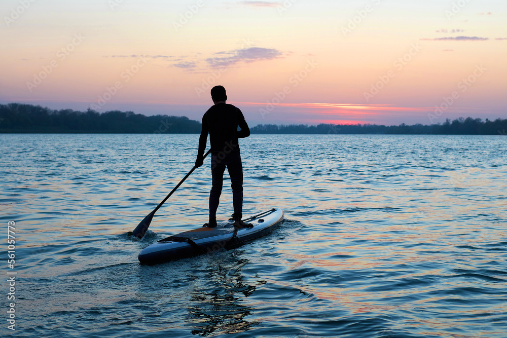 Silhouette of a man with a paddle on a SUP surfboard. Picturesque body of water in the evening. Theme of an active lifestyle