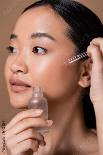 Young asian woman applying serum on cheek isolated on brown.