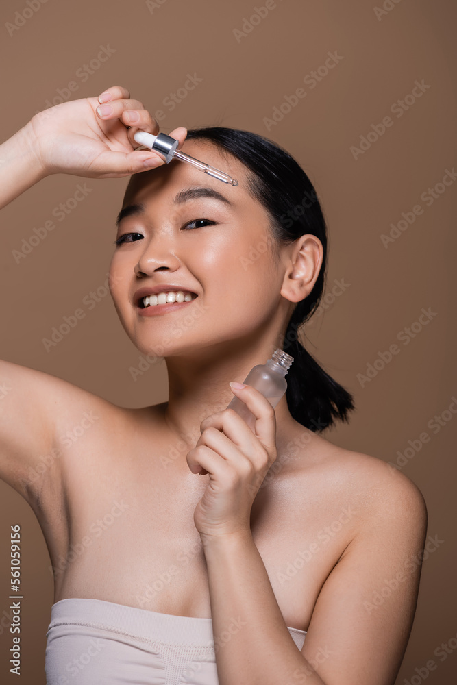 Φωτογραφία Pleased asian woman in top holding pipette and bottle of cosmetic serum isolated on brown