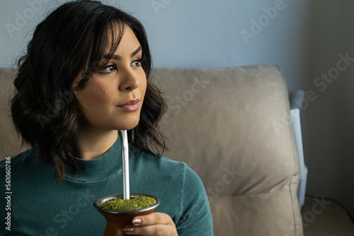 Mulher morena de cabelo curso enrolado vestindo uma blusa verde sentada no sofá tomando chimarrão photo