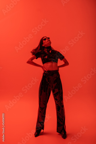 full length of stylish woman in latex crop top and trousers standing with hands on hips on red.