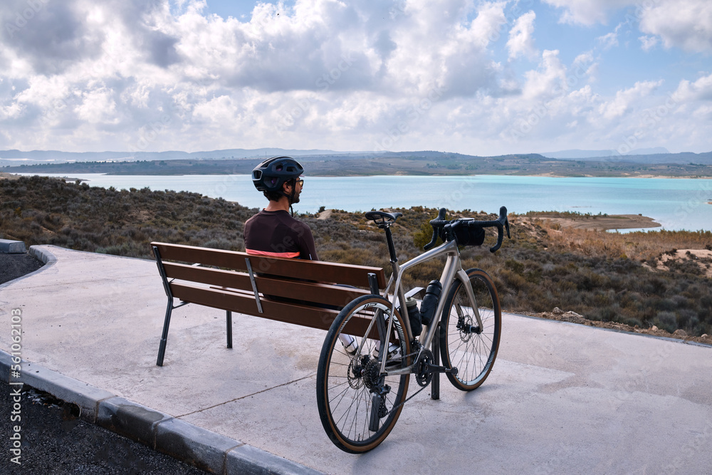 A male cyclist rests on a bench overlooking a blue lake and mountains .
Stylish handsome guy cyclist rests on a wooden bench near bicycle in summer park.