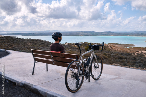 A male cyclist rests on a bench overlooking a blue lake and mountains . Stylish handsome guy cyclist rests on a wooden bench near bicycle in summer park.
