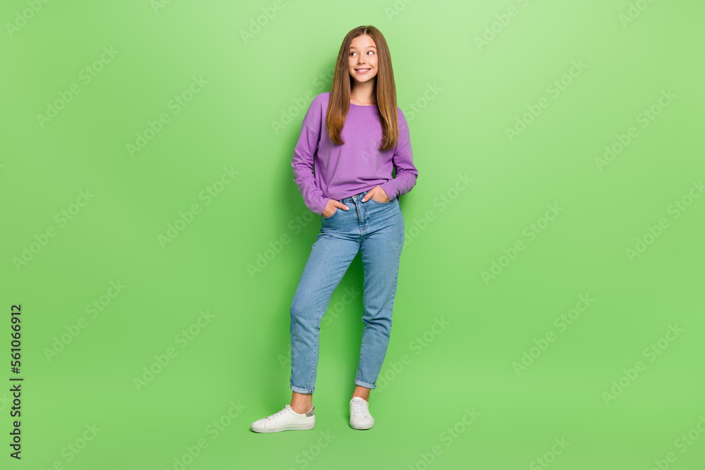 Full length photo of optimistic girl straight hairdo purple pullover arms in pockets look empty space isolated on green color background Full length photo of optimistic girl straight hairdo purple pullover arms in pockets look empty space isolated on green color background