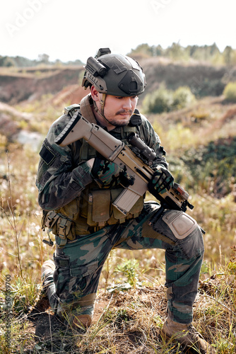 Brave male in combat uniform is standing in nature alone waiting for operation, strong soldier man in combat uniform holding rifle weapon in hands, posing.