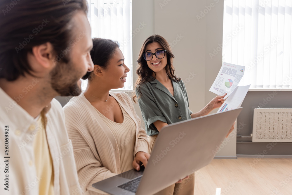 happy businesswoman in eyeglasses showing infographics to colleagues standing with laptop in office. happy businesswoman in eyeglasses showing infographics to colleagues standing with laptop in office.