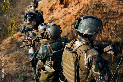 Squad of Young Athlete Fully Equipped Friendly Soldiers in Camouflage on a Reconnaissance Military Mission, Aiming Rifles. Rear View On Men Moving in Formation Through Mountains. © Roman