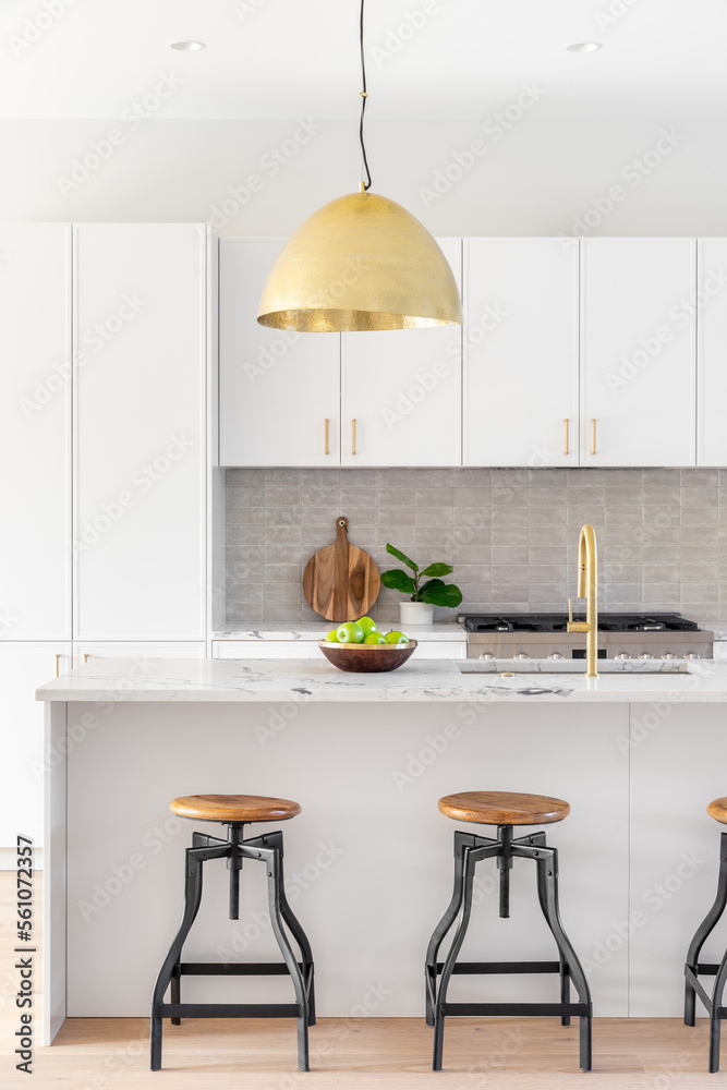 A kitchen detail with white cabinets, gold faucet and light hanging over  the island with bar stools, and a tiled backsplash. Stock Photo | Adobe  Stock