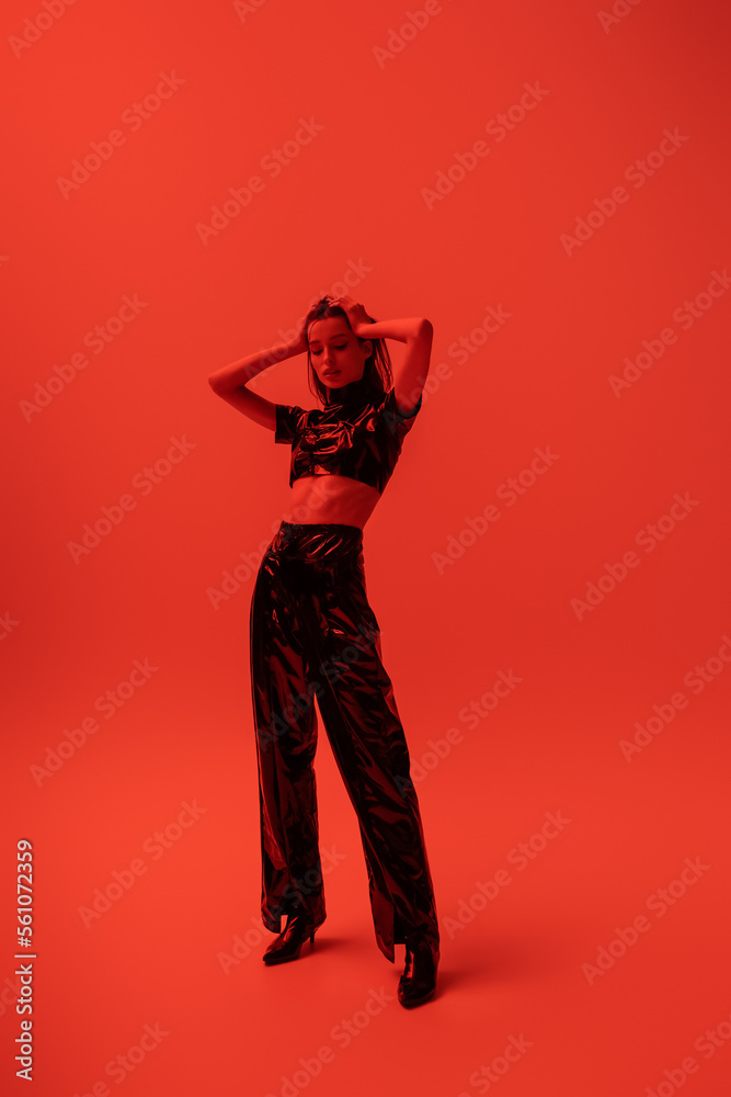 full length of stylish woman in latex crop top and trousers posing with hands on head on red. full length of stylish woman in latex crop top and trousers posing with hands on head on red.