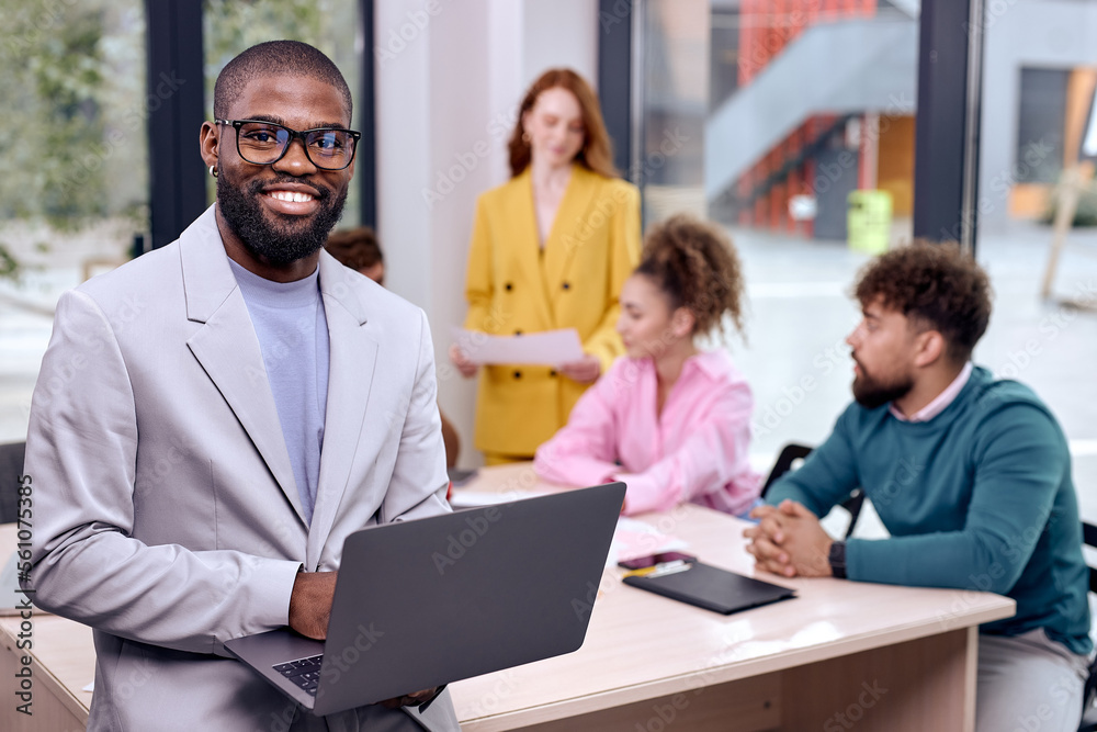 Portrait of happy black business man posing with laptop while colleagues working in background, diverse people gathered in modern office, cooperating, collaborating. success, people concept Portrait of happy black business man posing with laptop while colleagues working in background, diverse people gathered in modern office, cooperating, collaborating. success, people concept
