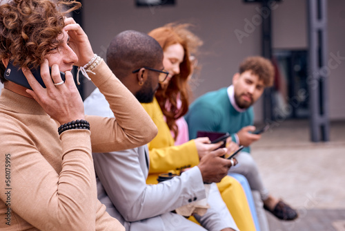 happy businesspeople walking, sitting on bench, using smartphones. young Caucasian and african colleagues chatting. focus on curly european guy talking on phone. outside of office. business people