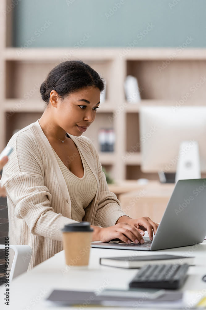 focused multiracial manager typing on laptop near blurred paper cup and notebook on work desk. focused multiracial manager typing on laptop near blurred paper cup and notebook on work desk.