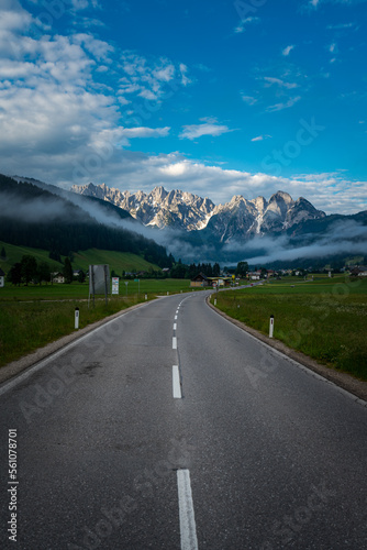 Straße in den Alpen mit Bergen, Wolken, grünen Wiesen und blauem Himmel.