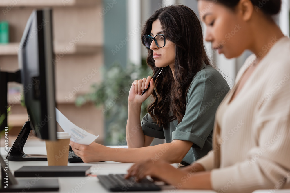 thoughtful businesswoman in eyeglasses looking at document near multiracial colleague typing on blurred foreground. thoughtful businesswoman in eyeglasses looking at document near multiracial colleague typing on blurred foreground.