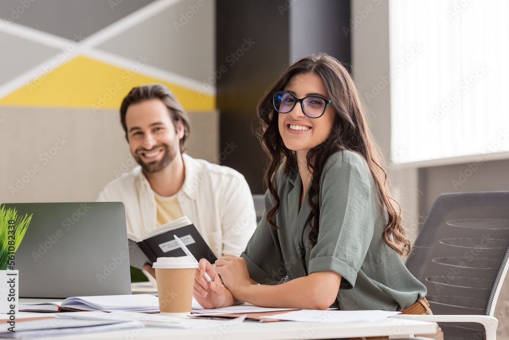 happy manager in eyeglasses smiling at camera near blurred colleague and paper cup on work desk. happy manager in eyeglasses smiling at camera near blurred colleague and paper cup on work desk.