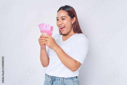 Excited young blonde woman girl Asian wearing casual white t-shirt looking at money rupiah banknotes in hand and rejoicing success isolated on white background. people lifestyle concept