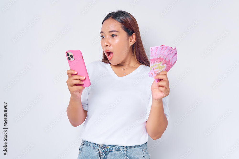 Shocked young blonde woman girl Asian wearing casual white t-shirt using mobile phone and holding money rupiah banknotes isolated on white background. Fast credits concept Shocked young blonde woman girl Asian wearing casual white t-shirt using mobile phone and holding money rupiah banknotes isolated on white background. Fast credits concept