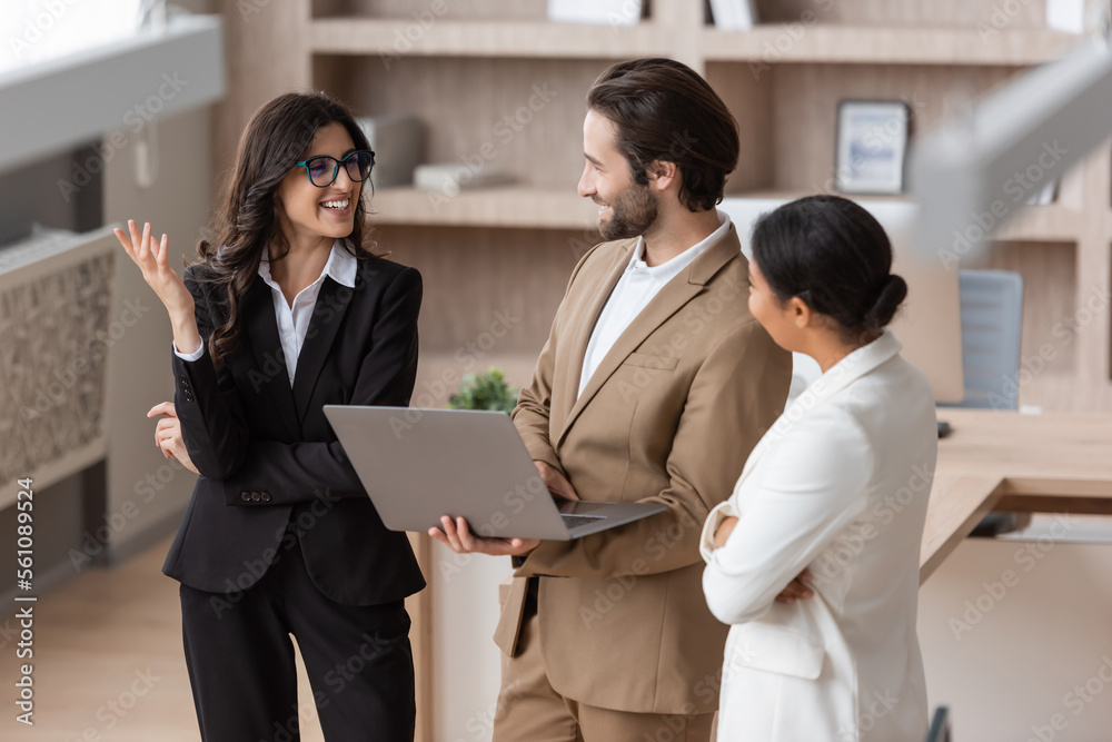 multiracial businesswoman and man with laptop looking at cheerful colleague in eyeglasses talking and gesturing in office. multiracial businesswoman and man with laptop looking at cheerful colleague in eyeglasses talking and gesturing in office.