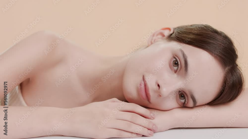 Medium shot of attractive young slim brown-haired Caucasian woman puts her head on her bent arm lying on white surface and smiles for the camera on beige background | Smooth arm skin concept
