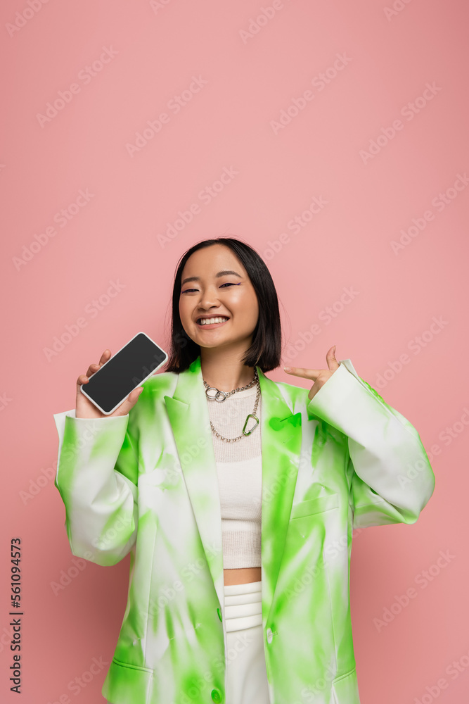 young asian woman in trendy jacket and necklaces pointing at smartphone with blank screen isolated on pink. young asian woman in trendy jacket and necklaces pointing at smartphone with blank screen isolated on pink.