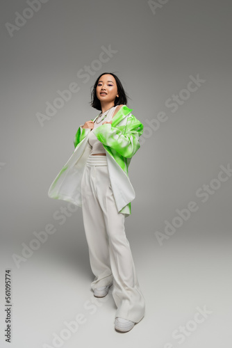 full length of young asian woman in pants and green and white jacket looking at camera on grey background.