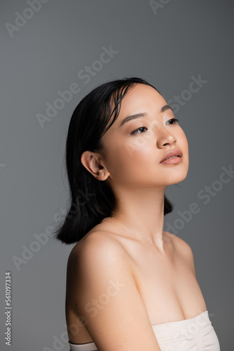 pretty asian woman with bare shoulders and nude makeup looking away isolated on grey.