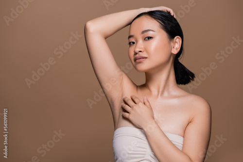 Pretty asian woman with naked shoulders touching fair and looking at camera isolated on brown.