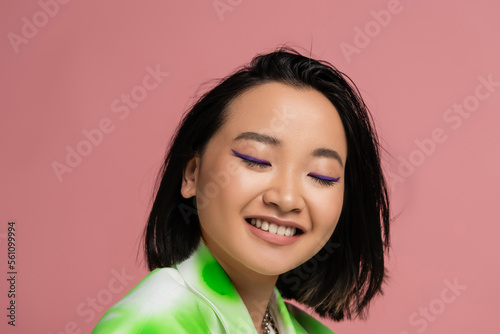 portrait of brunette asian woman with makeup smiling with closed eyes isolated on pink.