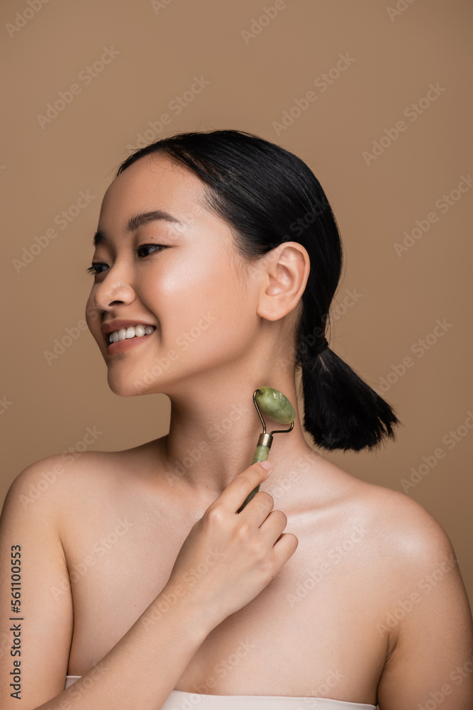 Happy asian woman with naked shoulders using jade roller on neck isolated on brown. Happy asian woman with naked shoulders using jade roller on neck isolated on brown.