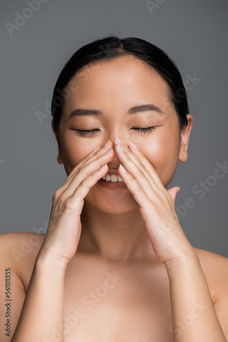 happy asian woman with closed eyes and perfect skin touching face isolated on grey.