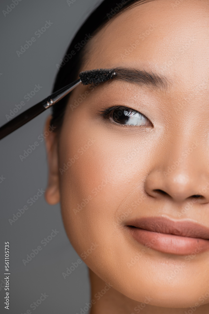 close up view of cropped asian woman with makeup foundation on face brushing eyebrow isolated on grey.