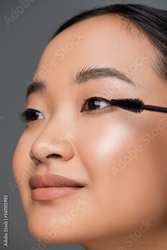 close up view of asian woman with makeup foundation applying black mascara isolated on grey.