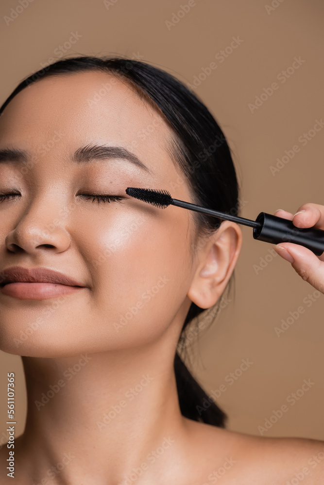 Cropped view of young asian woman holding applicator for mascara isolated on brown. Cropped view of young asian woman holding applicator for mascara isolated on brown.