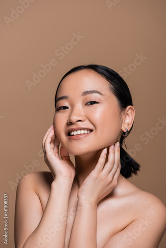 Young asian woman with naked shoulders touching checks isolated on brown.