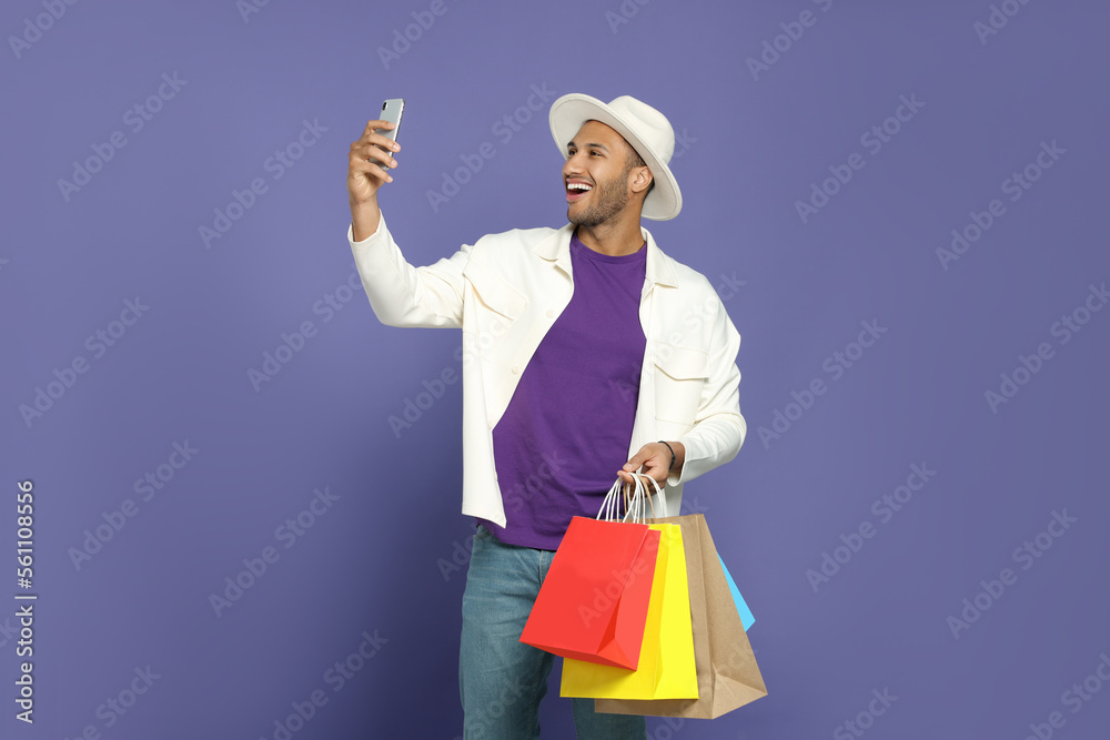 Happy African American man in hat with shopping bags taking selfie on purple background Happy African American man in hat with shopping bags taking selfie on purple background