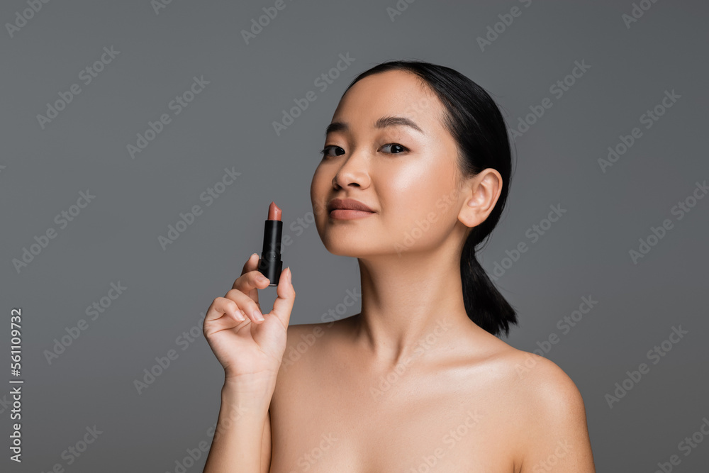 Pretty asian woman with naked shoulders holding beige lipstick isolated on grey. Pretty asian woman with naked shoulders holding beige lipstick isolated on grey.