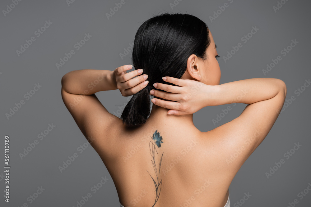 Brunette asian woman with tattoo on back touching ponytail isolated on grey. Brunette asian woman with tattoo on back touching ponytail isolated on grey.