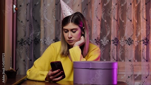 The unhappy girl stayed at home alone on her birthday, in a festive hat, flatters the news feed on the Internet, disappointment. Sad holidays. Social distancing. Isolation concept. photo