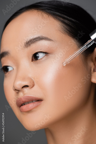 close up view of asian woman applying moisturizing cosmetic serum during facial pampering isolated on grey.