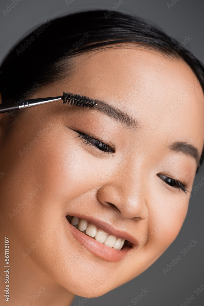 portrait of cheerful asian woman with natural makeup brushing eyebrow isolated on grey.