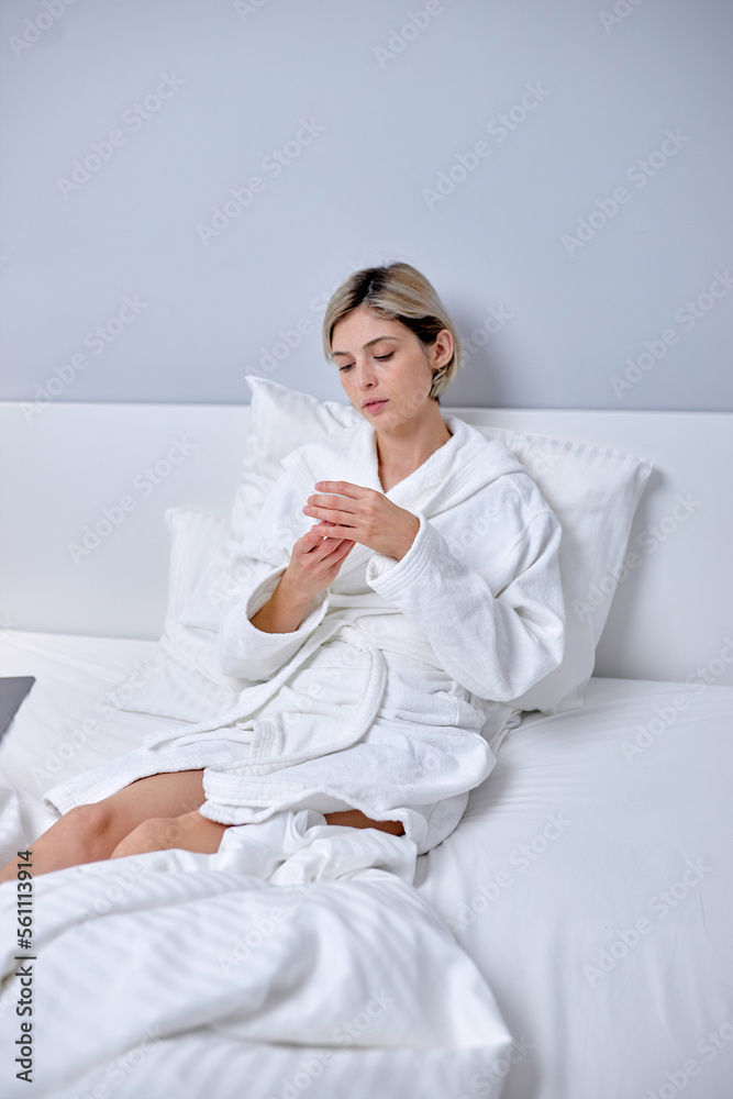 adorable female in bathrobe sitting on bed with cream bottle in hands, have rest, in the morning at weekends. caucasian european lady with short hair enjoy holidays alone at home in bedroom adorable female in bathrobe sitting on bed with cream bottle in hands, have rest, in the morning at weekends. caucasian european lady with short hair enjoy holidays alone at home in bedroom