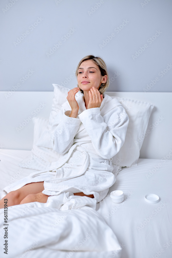 young female in bathrobe applying cream on face touching moisturized soft healthy sensitive skin doing morning hydration spa beauty routine sitting on bed, facial skincare treatment concept