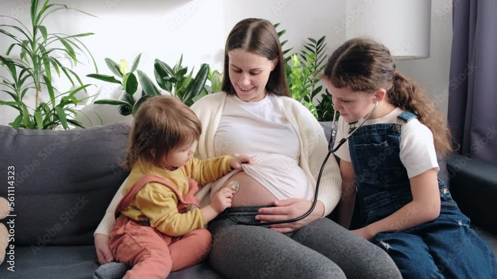 Cute two little daughters applying stethoscope to pregnant mom belly, listening to younger sibling motions in abdomen. Expecting mother playing with kids, having fun. Childhood and motherhood concept Cute two little daughters applying stethoscope to pregnant mom belly, listening to younger sibling motions in abdomen. Expecting mother playing with kids, having fun. Childhood and motherhood concept
