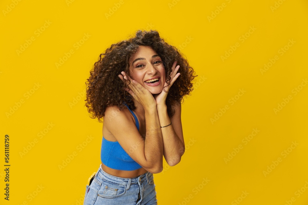 Woman with curly afro hair in a blue t-shirt on. yellow background signs with her hands, look into the camera, smile with teeth and happiness, copy space Woman with curly afro hair in a blue t-shirt on. yellow background signs with her hands, look into the camera, smile with teeth and happiness, copy space