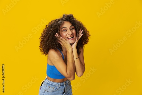 Woman with curly afro hair in a blue t-shirt on. yellow background signs with her hands, look into the camera, smile with teeth and happiness, copy space © SHOTPRIME STUDIO